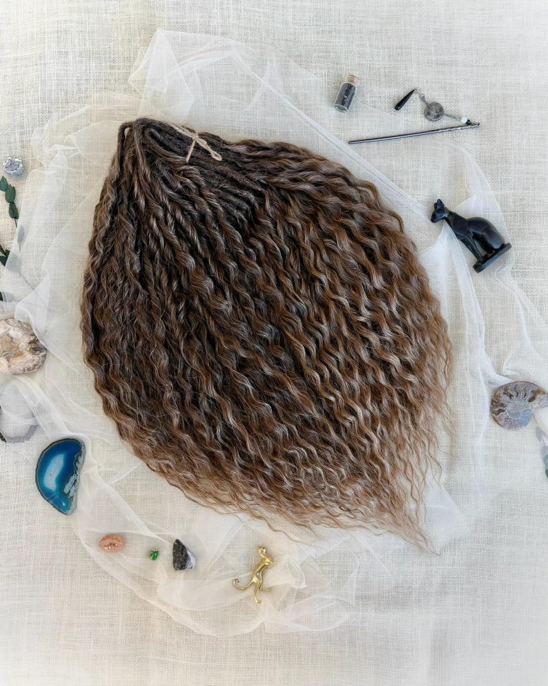 May include: A bundle of brown and blonde synthetic hair extensions with a wavy, twisted texture. The hair is arranged on a white fabric surface surrounded by small decorative items including a black cat figurine, stones, and a metal hair accessory.