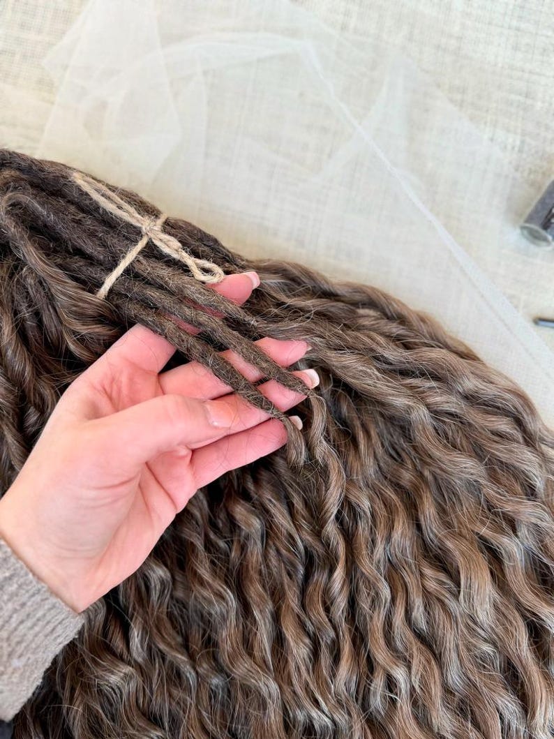 May include: A bundle of brown wavy dreadlocks tied together with a thin piece of twine. The hair extensions feature a multi-tonal brown color palette with lighter highlights throughout the wavy texture, creating a voluminous and layered appearance.