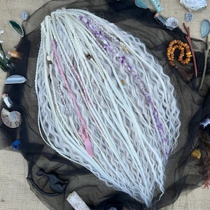 May include: White and light gray dreadlocks with pink and purple accents. The dreadlocks are arranged on a black mesh fabric, surrounded by decorative crystals, shells, and other small objects. The dreadlocks are wavy and have a natural appearance.