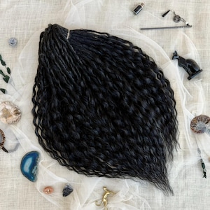 Puede incluir: Un paquete de rastas sintéticas negras con una textura ondulada. Las rastas están atadas con una banda marrón claro. La imagen también incluye elementos decorativos como una figura de gato negro, cristales y una pequeña botella.