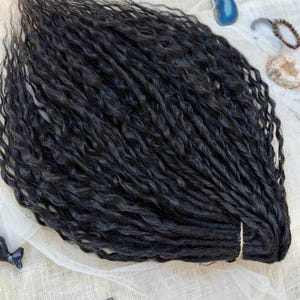 May include: A bundle of black synthetic hair extensions featuring a wavy, textured pattern. The individual strands are gathered and secured at one end with a light-colored band, creating a uniform, voluminous appearance with a consistent dark color.