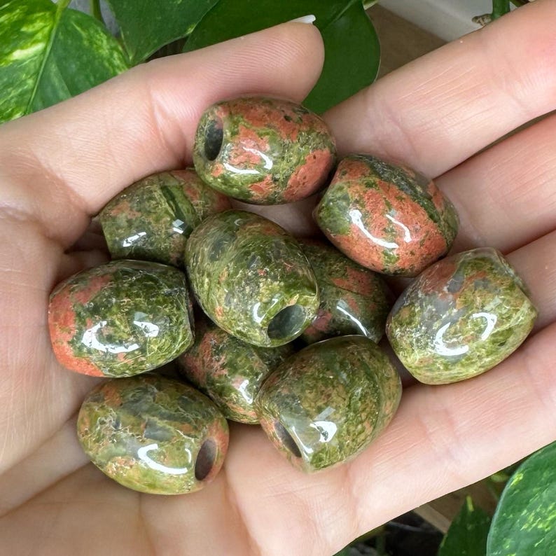 May include: A collection of polished, barrel-shaped beads made from unakite stone. Each bead features a mottled, smooth surface with a distinct blend of earthy green and salmon pink mineral inclusions, and a central hole drilled through the middle.