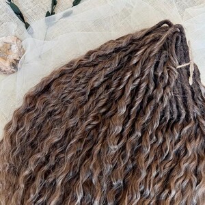 May include: A bundle of brown hair extensions featuring a wavy, twisted texture. The strands exhibit a multi-tonal brown color palette with lighter highlights throughout, secured at the top with a thin, dark brown cord tied in a small knot.