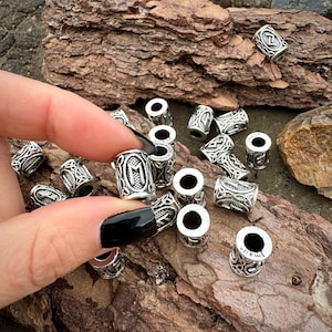 May include: Silver-colored metal beads with intricate Celtic knot designs and runes. The beads are cylindrical with a hole running through the center. Some beads are held by a person with black nail polish. The beads are scattered on a brown, textured surface.