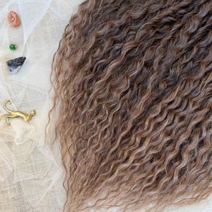 May include: A section of long, wavy, light brown hair extensions with subtle highlights. The hair is arranged next to a small gold-colored cat figurine, a blue agate slice, a small pink stone, a green bead, and a dark grey stone on white fabric.