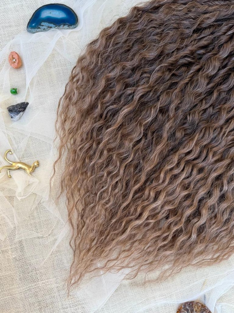 May include: A section of long, wavy, light brown hair extensions with subtle highlights. The hair is arranged next to a small gold-colored cat figurine, a blue agate slice, a small pink stone, a green bead, and a dark grey stone on white fabric.