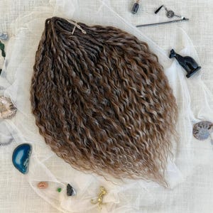 May include: A bundle of wavy, brown synthetic hair extensions with lighter highlights, arranged in a fan shape. The hair features a crimped, textured appearance with a central tie. Surrounding the hair are small decorative items including stones, a black cat figurine, a metal hair stick, and a small vial.