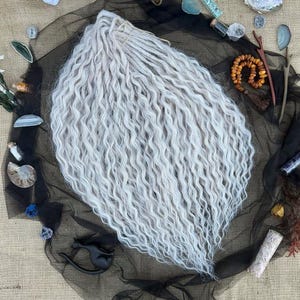 May include: A bundle of light gray, wavy synthetic dreadlocks. The dreadlocks are arranged on a black mesh fabric, surrounded by decorative crystals, stones, and small objects. The background is a textured, tan fabric.