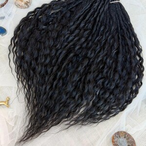May include: A bundle of black hair extensions featuring a wavy, textured pattern. The strands are gathered at the top with a light-colored string, creating a fan-like shape that cascades downward with defined, crimped curls throughout the length.