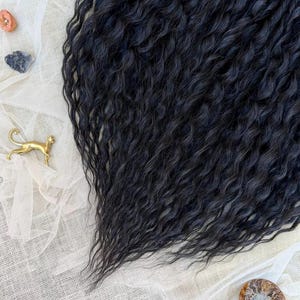 May include: A collection of long, wavy black hair extensions with a crimped texture. The set includes a small blue agate slice, a tiny orange stone, a dark grey stone, a gold-toned metal cat figurine, and a spiral ammonite fossil shell.
