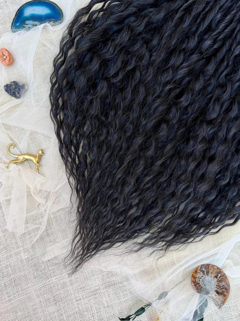 May include: A collection of long, wavy black hair extensions with a crimped texture. The set includes a small blue agate slice, a tiny orange stone, a dark grey stone, a gold-toned metal cat figurine, and a spiral ammonite fossil shell.