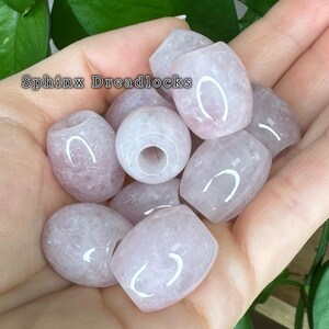 May include: A collection of pale pink, translucent, barrel-shaped beads with a smooth, polished finish and a central hole. The text 'Sphinx Dreadlocks' is visible in the upper left corner of the image, overlaying the light pink stone beads.