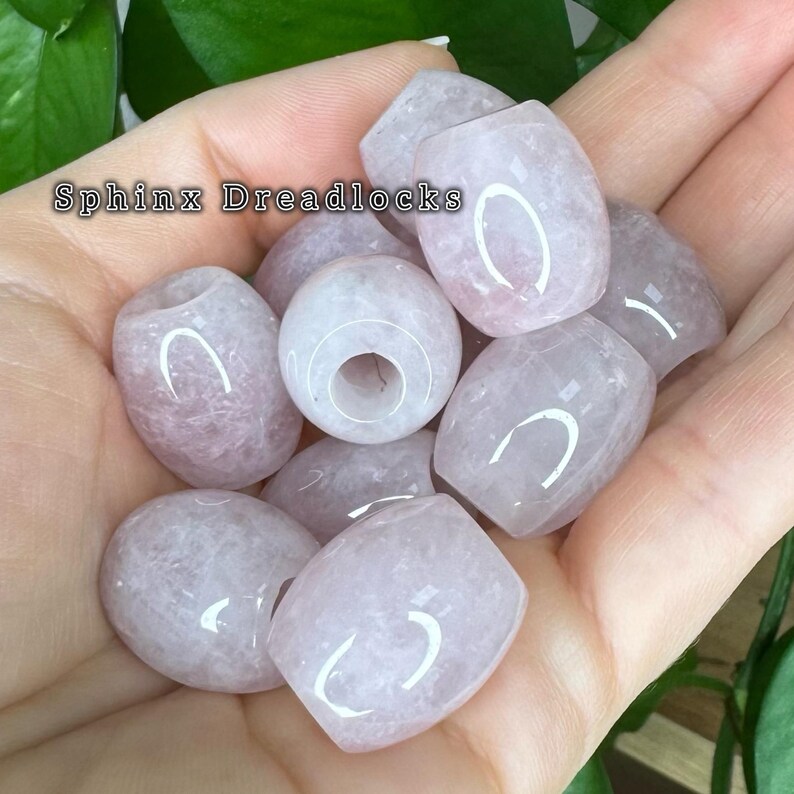 May include: A collection of pale pink, translucent, barrel-shaped beads with a smooth, polished finish and a central hole. The text 'Sphinx Dreadlocks' is visible in the upper left corner of the image, overlaying the light pink stone beads.