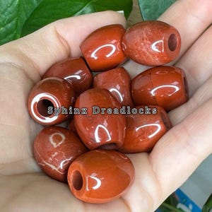 May include: A collection of smooth, polished, barrel-shaped red jasper beads with a central hole, featuring natural stone variations and glossy surfaces. The text "Sphinx Dreadlocks" is overlaid in the center of the image.