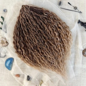 May include: A bundle of wavy, ombre hair extensions, transitioning from dark brown to a light blonde. The extensions are arranged on a white, sheer fabric, with decorative stones and a small gold figurine scattered around.