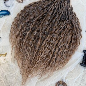 May include: A bundle of synthetic hair extensions featuring a wavy, crimped texture in a multi-tonal brown color palette. The strands transition from darker brown roots to lighter, golden-brown tips, arranged in a fan shape with a tied top section.