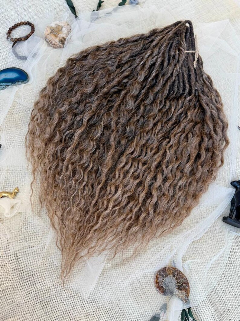 May include: A bundle of synthetic hair extensions featuring a wavy, crimped texture in a multi-tonal brown color palette. The strands transition from darker brown roots to lighter, golden-brown tips, arranged in a fan shape with a tied top section.