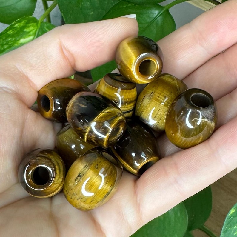 May include: A collection of polished tiger's eye gemstone beads, each featuring a smooth, barrel-shaped design with a central hole. The beads display natural golden-brown and dark brown chatoyant bands with a reflective, glossy finish.