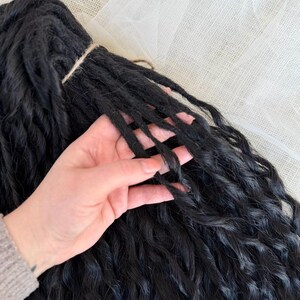 May include: A bundle of black synthetic hair extensions featuring a wavy, textured finish. The individual strands are gathered at the top with a thin cord, showcasing a consistent dark color and a soft, crimped appearance throughout the length.