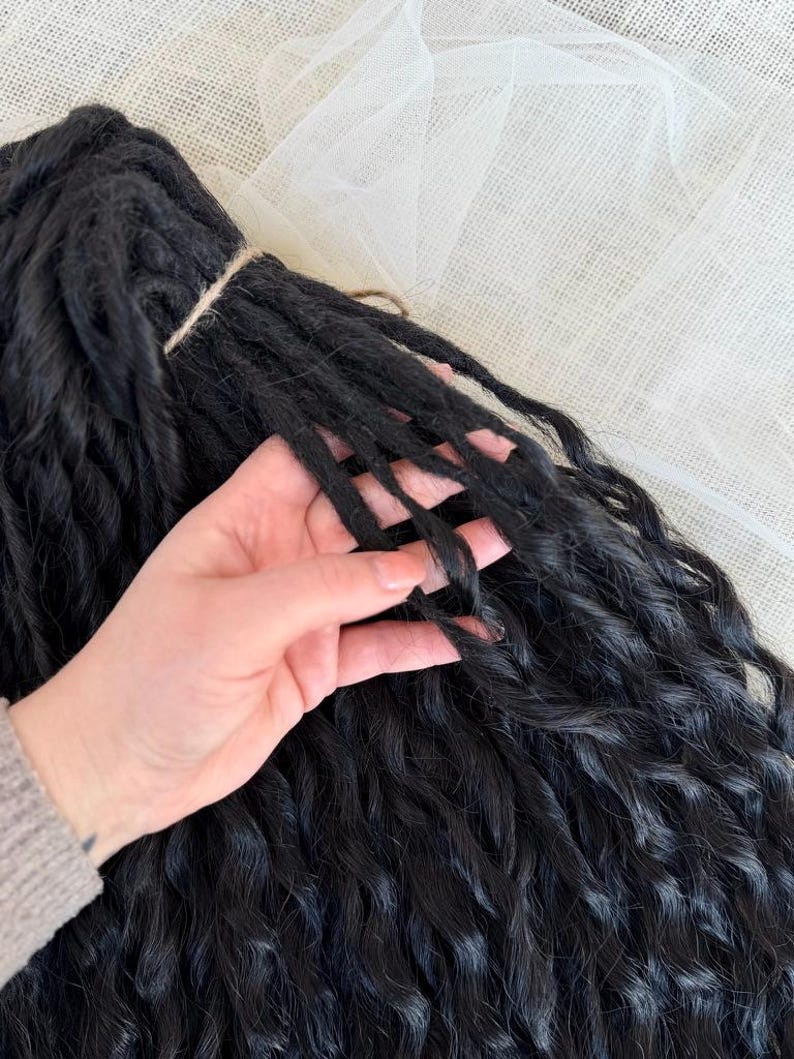 May include: A bundle of black synthetic hair extensions featuring a wavy, textured finish. The individual strands are gathered at the top with a thin cord, showcasing a consistent dark color and a soft, crimped appearance throughout the length.