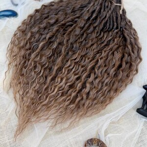 May include: A bundle of synthetic hair extensions featuring a dark brown to light brown ombre color gradient. The hair has a tight, wavy, and crimped texture throughout, with individual strands gathered and tied together at the top section.