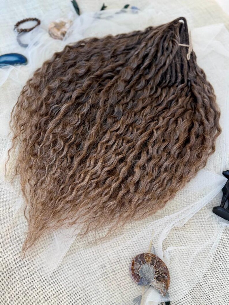May include: A bundle of synthetic hair extensions featuring a dark brown to light brown ombre color gradient. The hair has a tight, wavy, and crimped texture throughout, with individual strands gathered and tied together at the top section.