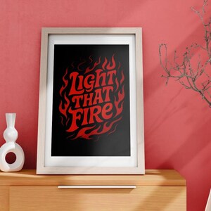 Puede incluir: Impresión artística enmarcada con las palabras "Light That Fire" en rojo, rodeadas de llamas, sobre un fondo negro. La obra de arte se exhibe en un mueble de madera con jarrones blancos y un jarrón alto con ramas.