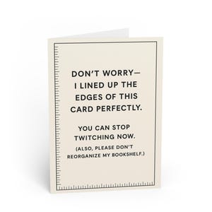 May include: A cream-coloured greeting card with black text that reads "DON'T WORRY - I LINED UP THE EDGES OF THIS CARD PERFECTLY. YOU CAN STOP TWITCHING NOW. (ALSO, PLEASE DON'T REORGANIZE MY BOOKSHELF.)". A black ruler design is on the left side.