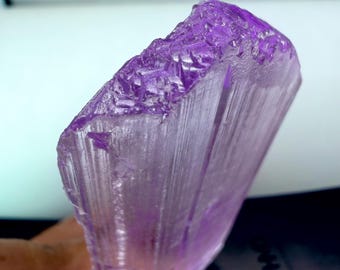 May include: A translucent, purple crystal with a rough, textured top and a smooth, striated body. The crystal is held in a hand, showcasing its natural form and color variations. The crystal's color ranges from light lavender to deep violet.