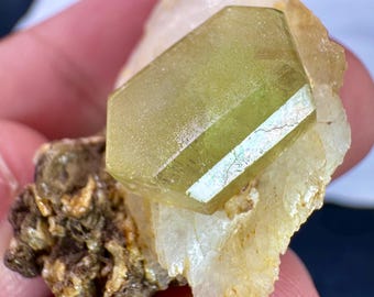 May include: A light green, hexagonal crystal with a reflective surface is set on a matrix of white and brown minerals. The crystal's facets catch the light, showcasing its clarity. The mineral specimen is held in a hand, highlighting its size and detail.