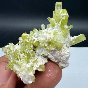 Peut inclure: Un amas de cristaux vert clair, possiblement un spécimen minéral, est posé sur une matrice blanche et texturée. Les cristaux sont allongés et translucides, avec une teinte vert-jaunâtre vibrante. La composition générale est naturelle et organique.