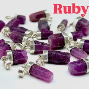 May include: A collection of raw, deep pinkish-red ruby pendants, each featuring a rough, unpolished gemstone secured in a silver-toned metal bail. The word Ruby is printed in red text at the top right of the image, which displays multiple pieces.