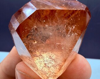 Op de afbeelding: Een gefacetteerd, roodbruin kristal met een diamantachtige vorm. Het kristal heeft een doorschijnend uiterlijk met interne insluitsels en wordt in een hand gehouden. Het oppervlak van het kristal reflecteert licht en toont zijn geometrische structuur.