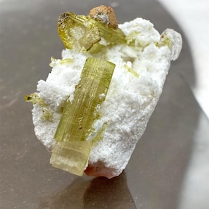 Puede incluir: Primer plano de un espécimen mineral con cristales de turmalina verde claro. Los cristales están incrustados en una matriz blanca y texturizada. También hay una pequeña formación marrón y terrosa.