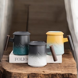 May include: Three ceramic mugs with lids and wooden handles. The mugs feature a mountain design in shades of black, teal, and yellow. The mugs are sitting on a white book that says "FOLIO".