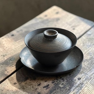 May include: A dark charcoal gray ceramic tea bowl with a matching lid and a circular saucer. The bowl features a smooth, matte finish with subtle variations in tone, a wide rim, and a small knob on the center of the lid for easy handling.