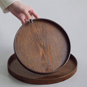 May include: A circular wooden tray with a dark brown finish and a visible natural wood grain pattern. The tray features a raised rim around the edge, providing a defined border for the flat surface. The wood displays deep brown tones.