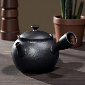 May include: A black ceramic teapot with a unique design. The teapot has a round body, a small spout, and a large, cylindrical handle. The lid has a small knob. The teapot is sitting on a wooden surface.