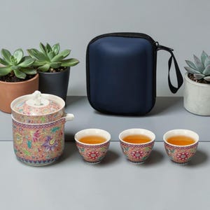 May include: A tea set with a floral design, including a teapot, three teacups, and a navy blue carrying case. The teapot and cups have a colorful, intricate pattern. Succulent plants in pots are in the background.