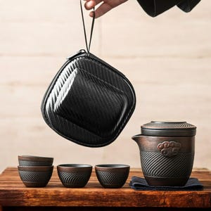 May include: A black, zippered carrying case is suspended above a tea set on a wooden table. The tea set includes a teapot with a lid, three small cups, and a small, dark blue cloth. The teapot and cups have a textured, dark brown finish.