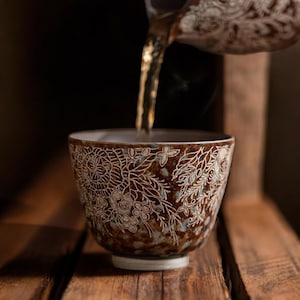 May include: A brown ceramic tea cup with a white floral design is being filled with tea. The cup has a wide mouth and a small, white base. The tea is a golden color, and steam is rising from the cup. The background is dark and out of focus.