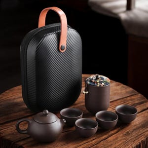 May include: A black tea set with a carrying case, teapot, cups, and a tea container. The case has a carbon fiber texture and a brown handle. The tea set is brown and sits on a wooden table.