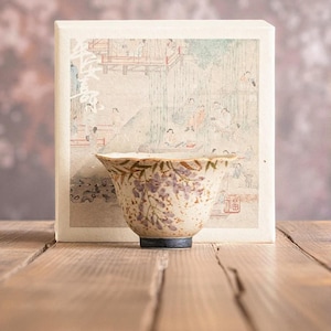 May include: A ceramic teacup with a floral design in purple and brown, set against a box with a traditional Japanese print. The cup has a dark blue base and sits on a wooden surface.