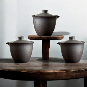 May include: Three dark brown ceramic tea sets with lids. One set is on a small wooden stand, and two are on a round wooden table. The tea sets have a textured, speckled finish and a spout for pouring.