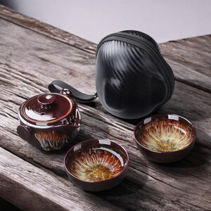 May include: A tea set comprising a dark brown glazed teapot, two matching cups, and a black carrying case. The cups and teapot feature a unique, colourful interior design. The set is arranged on a rustic wooden surface.