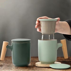 Puede incluir: Dos tazas de té de cerámica con asas de madera. Una es verde oscuro con tapa, la otra es verde claro con un infusor de té. Una mano vierte líquido del infusor en la taza verde claro. Las tazas están sobre un posavasos de madera.