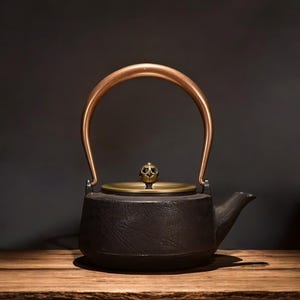 May include: A black cast iron teapot with a copper handle and lid. The teapot has a spout and sits on a wooden surface. The lid is gold-colored and has a decorative knob.