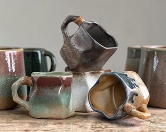May include: A collection of handmade ceramic mugs in various shapes and colors. The mugs feature unique designs, including angular forms and textured surfaces. Some mugs have handles wrapped with a natural fiber. The color palette includes earthy tones like brown, green, and blue.