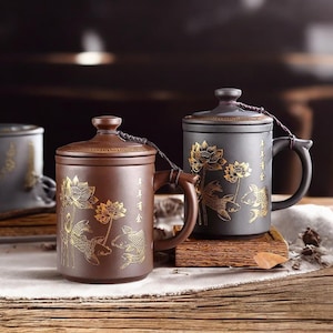 May include: Two ceramic tea mugs with lids. One is brown, the other is dark grey. Both mugs feature gold lotus flowers and koi fish designs. The mugs have handles and are on a wooden surface.