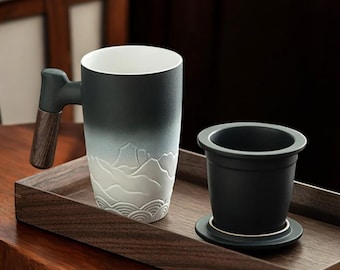May include: A ceramic tea mug with a gradient dark green to white design, featuring a mountain landscape relief. It has a wooden handle and sits on a wooden tray, alongside a black tea infuser.